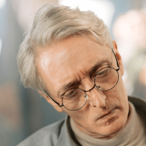 A middle-aged man with gray hair and glasses looks downward with a sad expression.