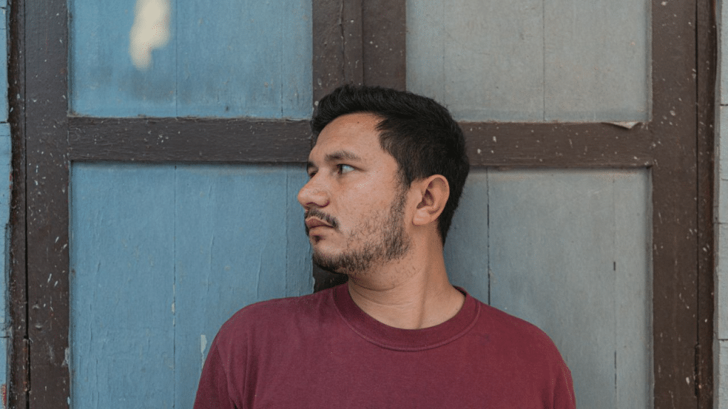 A man with a goatee and a maroon shirt looks off to the side, standing in front of a blue and brown wooden door.