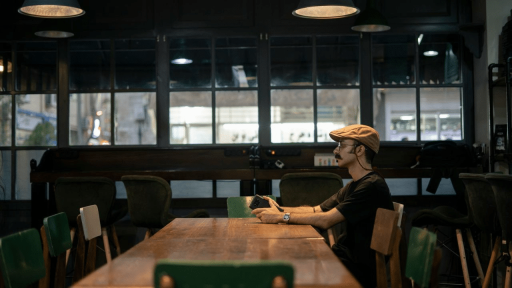 A man wearing a flat cap sits at a long wooden table in a dimly lit restaurant.