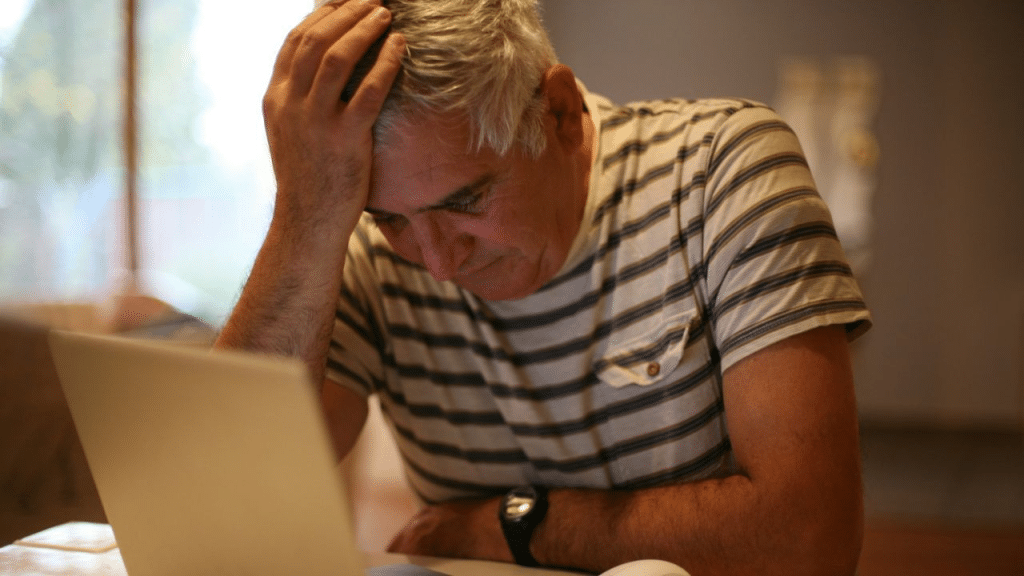 A middle-aged man with gray hair in a striped shirt sits at a desk, holding his head in his hand and looking at a laptop.