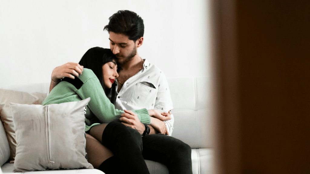 A man and woman sit together on a white couch; the man is hugging the woman, who is resting her head on his chest.