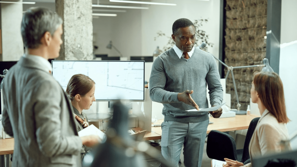 A diverse group of four office workers stands and sits, discussing papers while looking at each other.