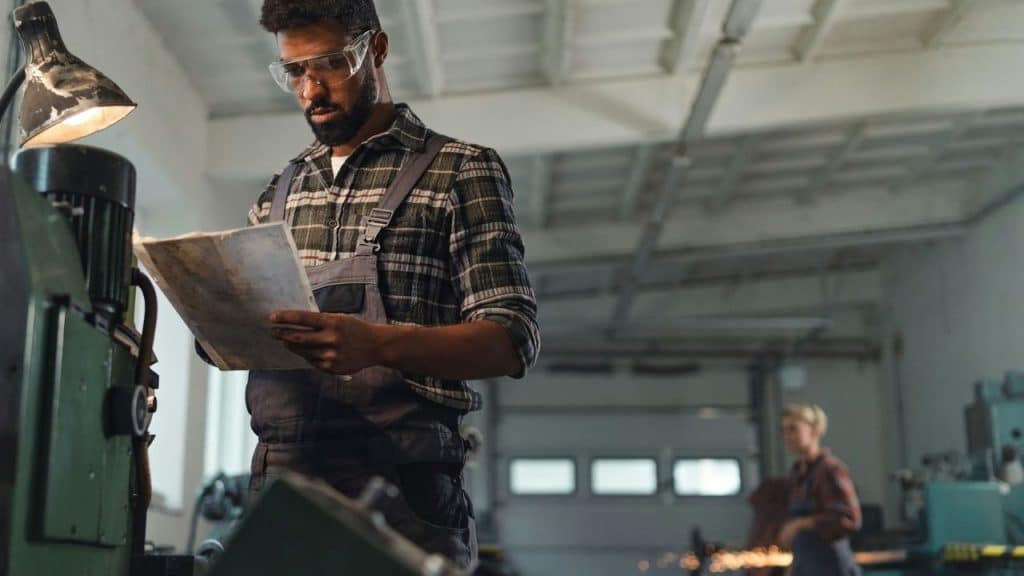 A bearded man with safety glasses and overalls works in a workshop.