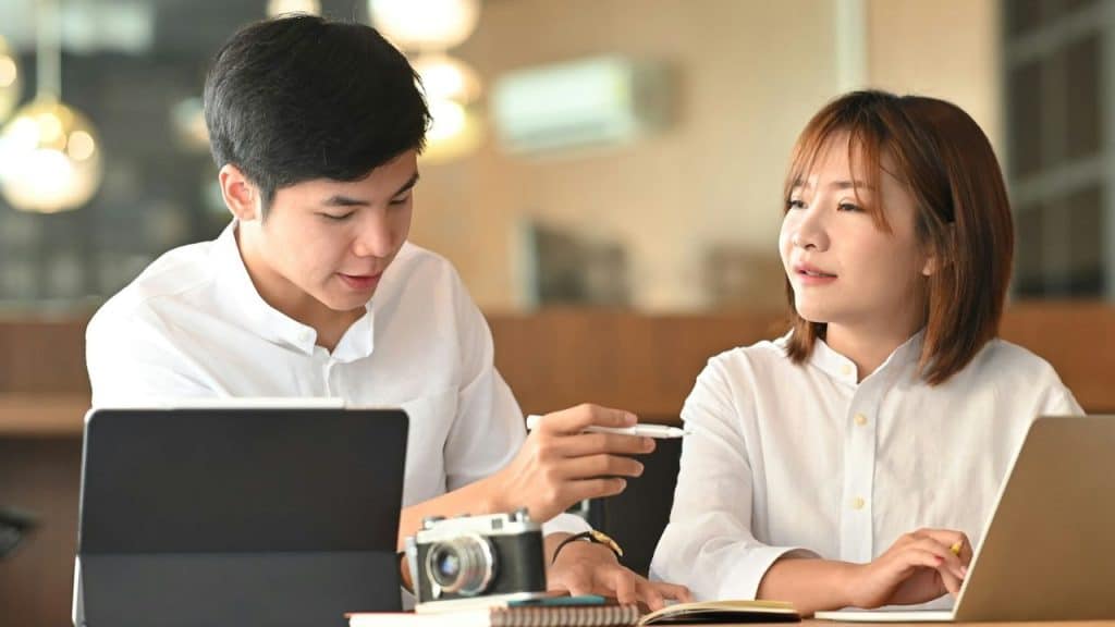A young man and woman sit together at a table with laptops, talking.