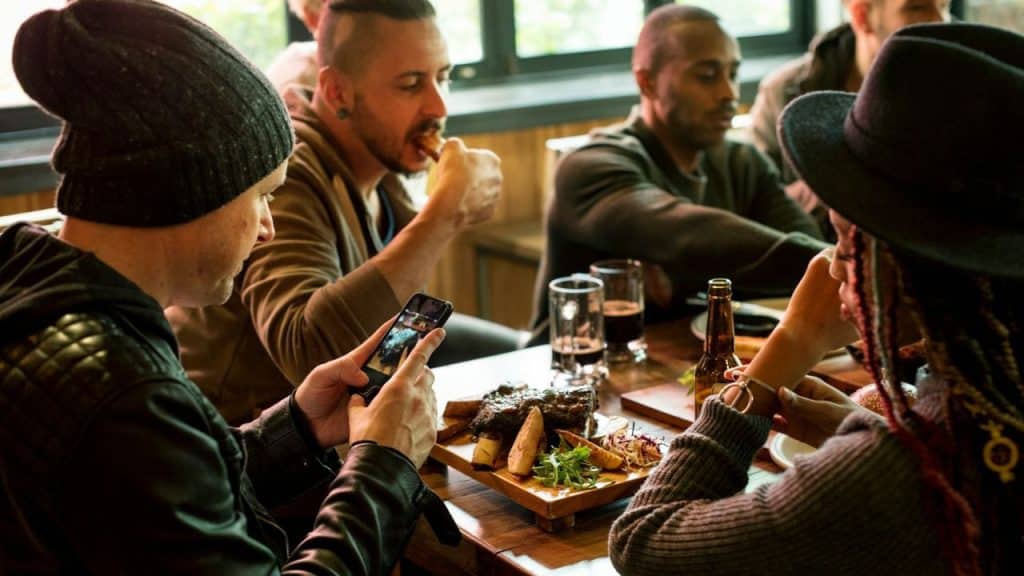 A group of people sits at a table, with one man taking a photo of food.