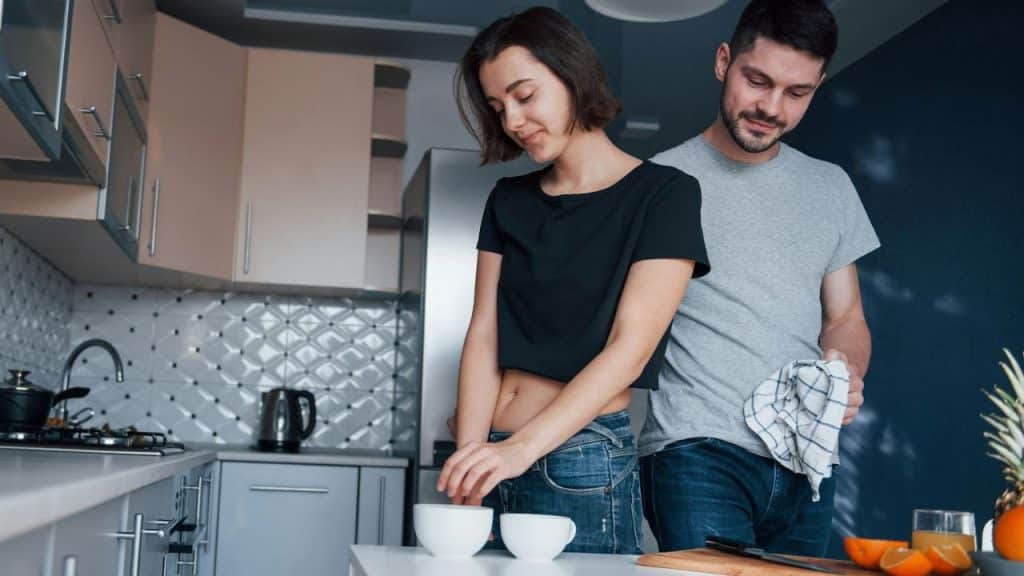 A smiling couple stands together in a kitchen, holding cups and a towel.