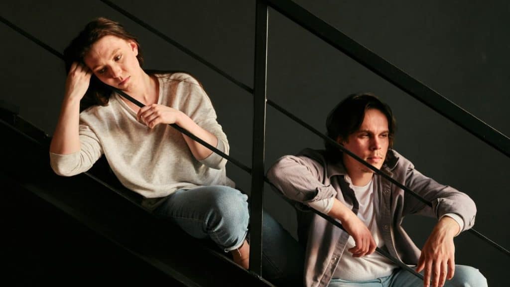A solemn man and woman sit on a staircase, looking away from each other.