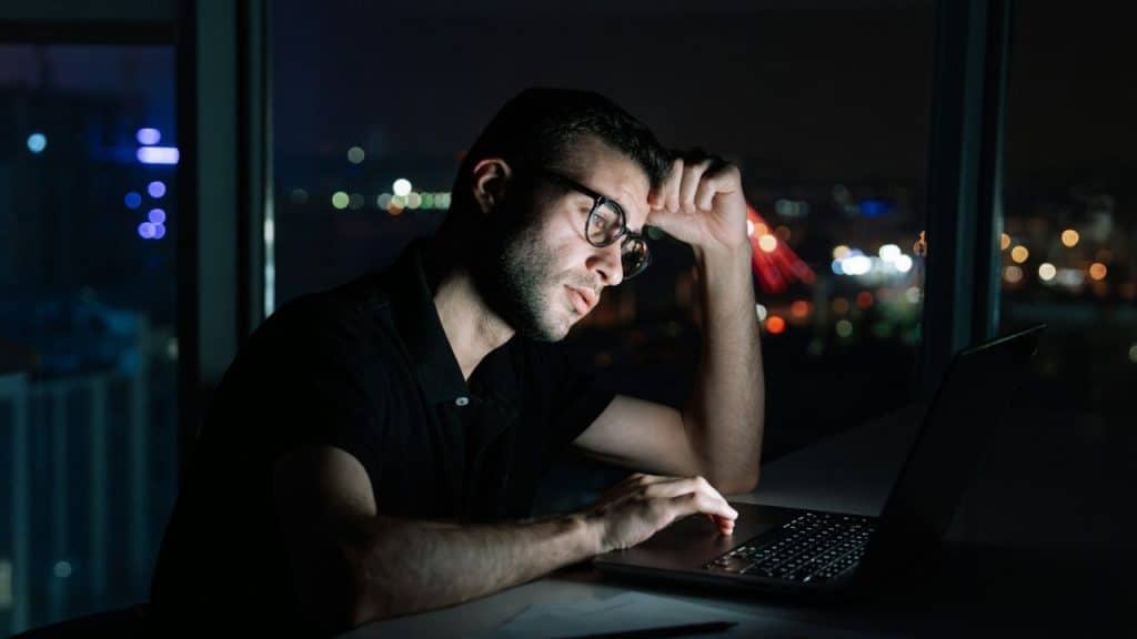 A serious man in glasses sits at a desk, working late on a laptop.