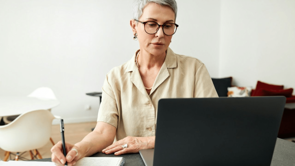 A focused woman with short gray hair and glasses writes in a notebook.
