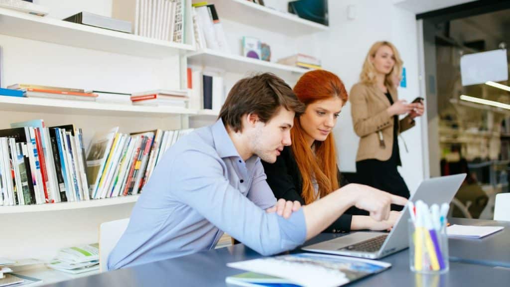 A man and woman sit at a desk, with the man pointing at a laptop screen.