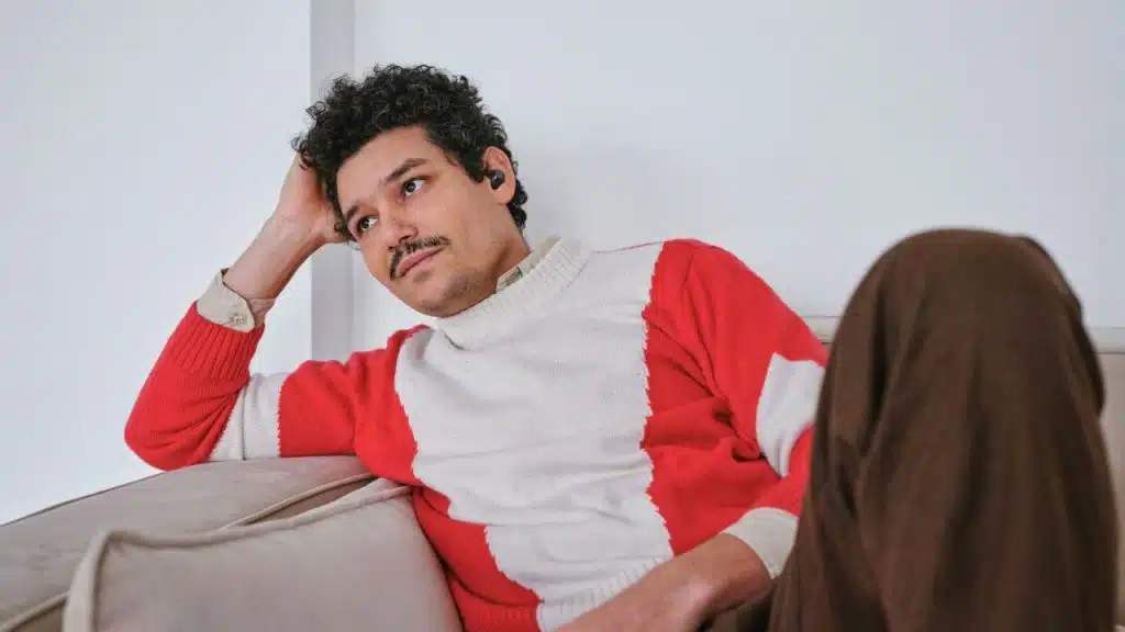 A man with a mustache and curly hair leans back on a couch, looking pensive.