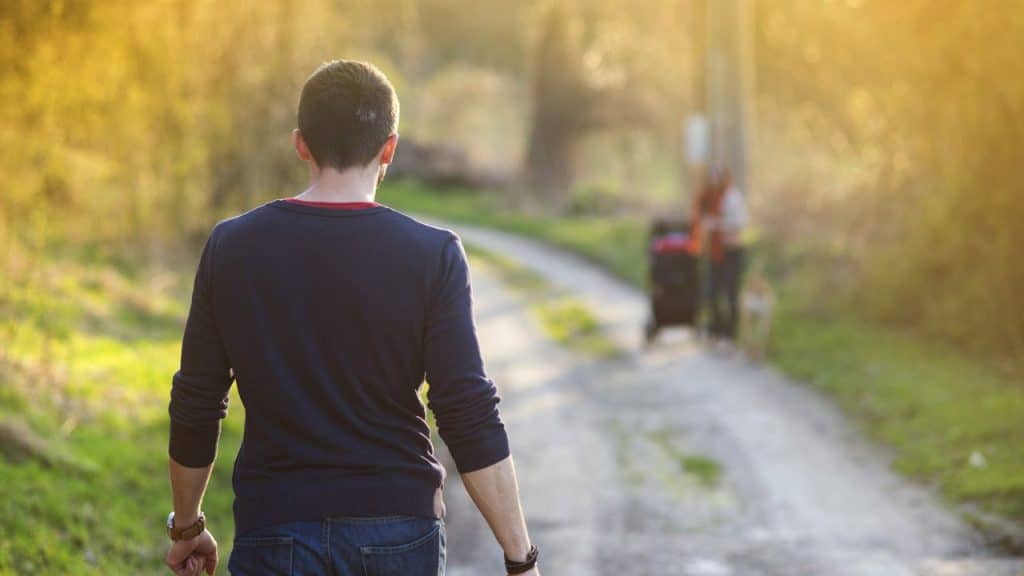 A man's back is to the camera as he walks down a dirt path, away from people in the distance.