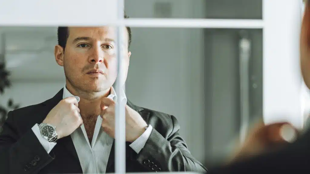 A man in a suit looks in a mirror as he adjusts his shirt collar.