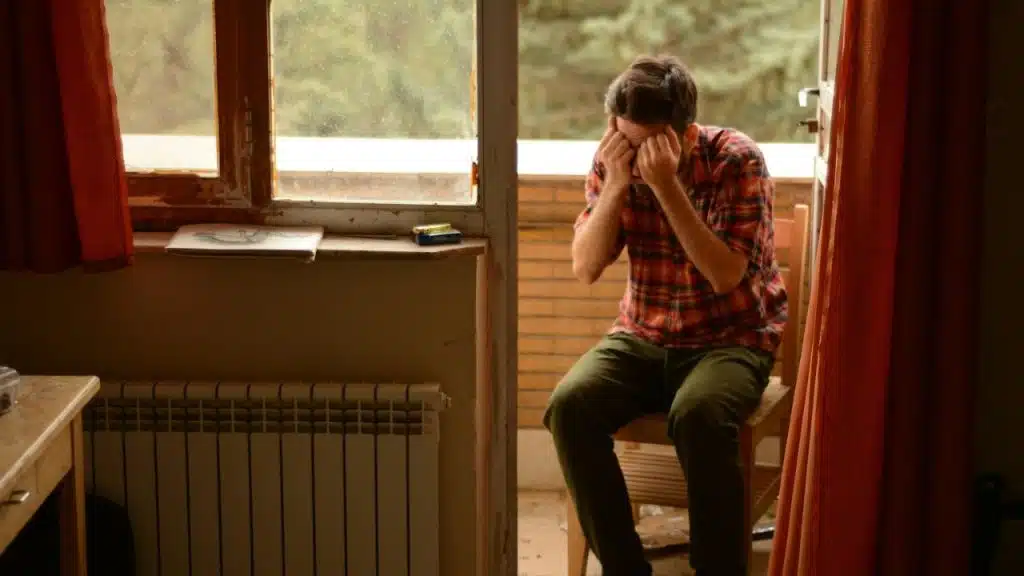 A man in a plaid shirt sits on a chair on a balcony, with his hands over his face.