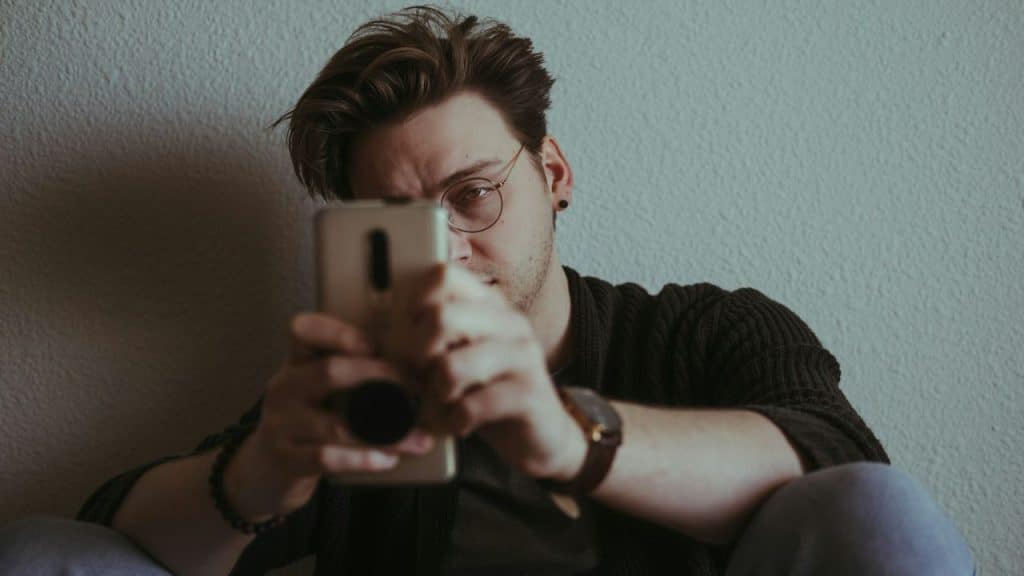A man wearing glasses looks at a phone he's holding with both hands.