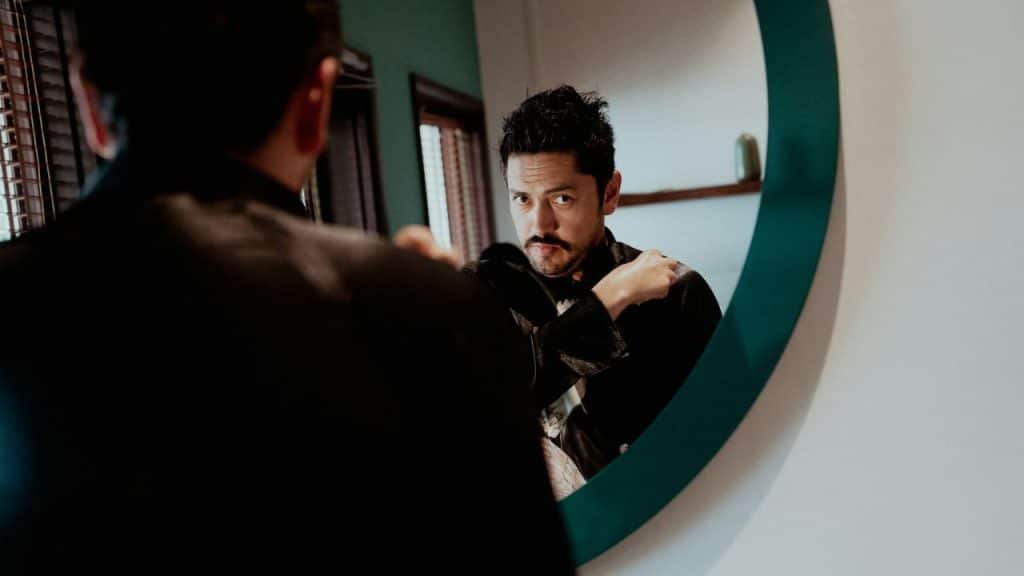 A man with a mustache looks at his reflection in a round mirror.