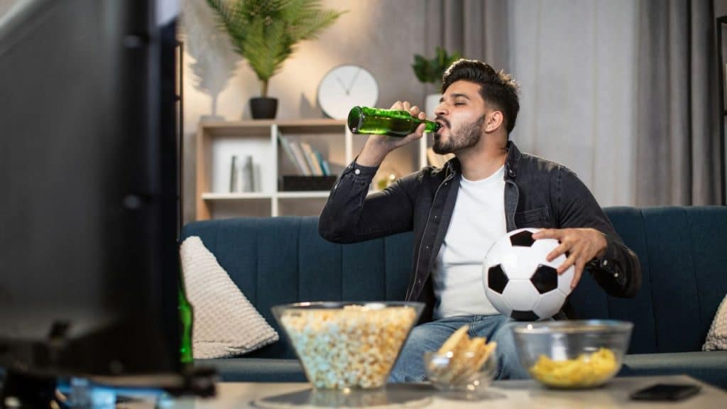 A man on a couch drinks from a green bottle, holding a soccer ball. Bowls of snacks sit on a table.