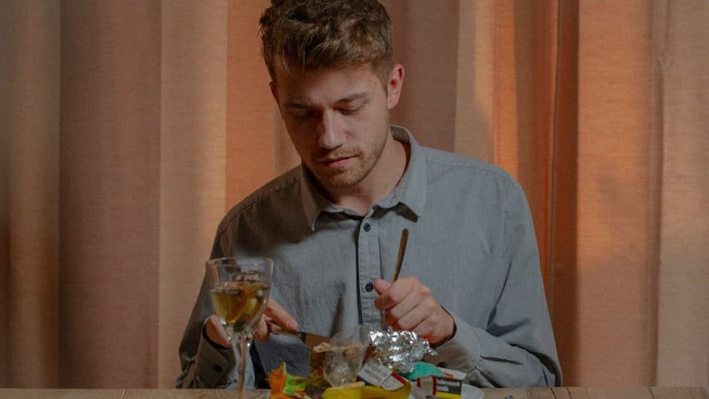 A man sits alone at a table with junk food wrappers and a glass of wine, looking down at his food.