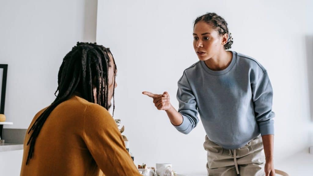 A woman stands, pointing her finger at a person with dreadlocks sitting at a table.