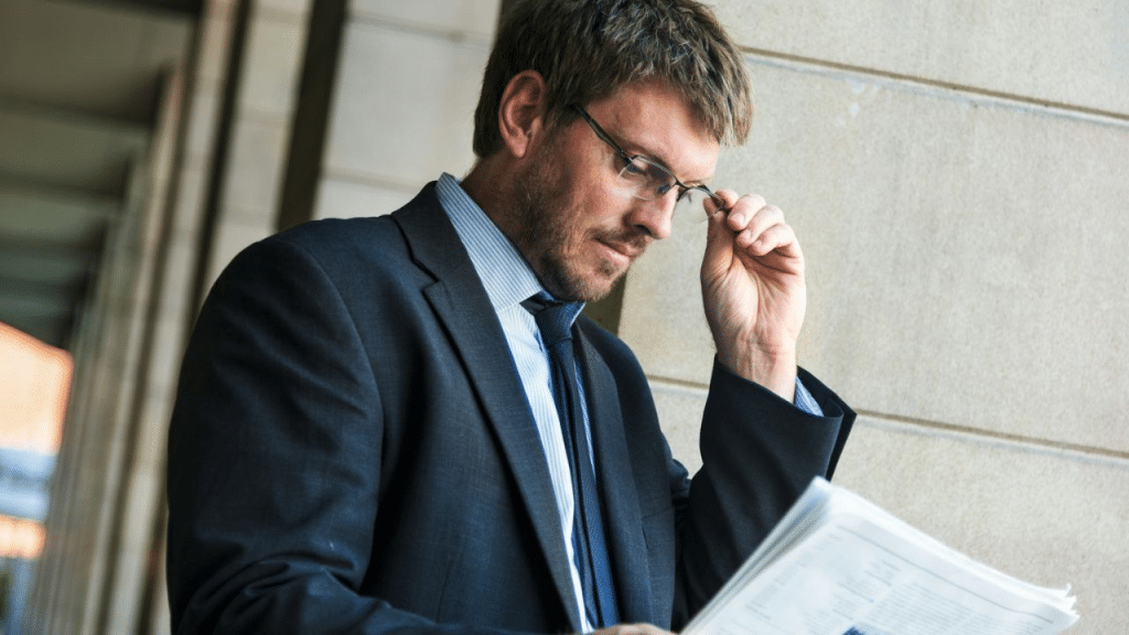 A man in a suit and tie stands outdoors, adjusting his glasses and reading a newspaper.