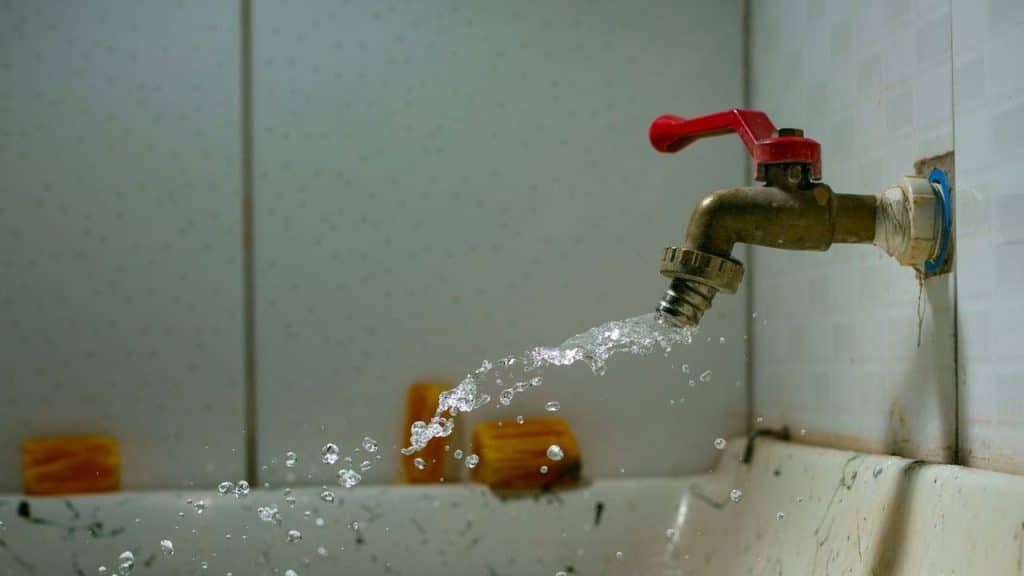 Water splashes from a faucet with a red handle into a dirty basin.