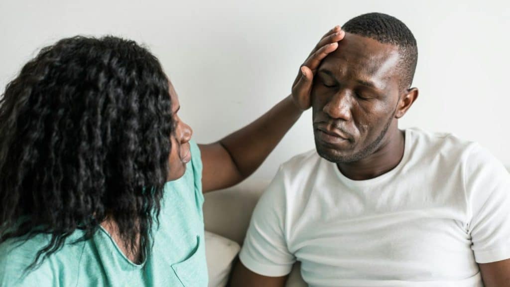 A woman is touching the forehead of a man who looks sick, with his eyes closed.