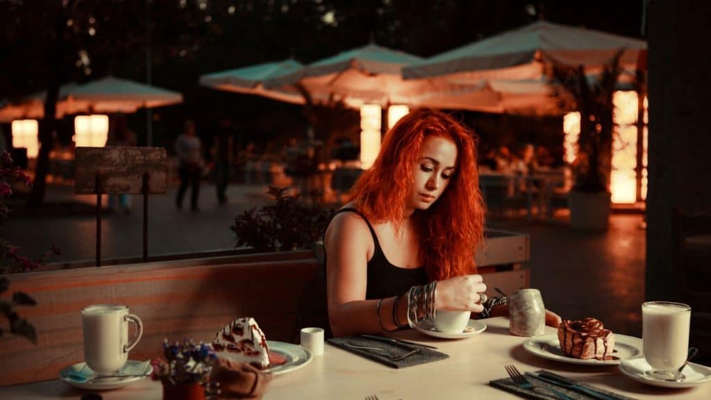 A woman with red hair sits alone at an outdoor cafe table, looking down at her cup.