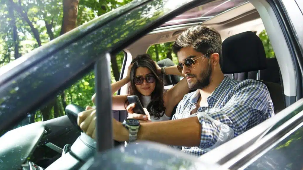A man is looking at his phone in the driver's seat, while a woman in the passenger seat holds her head in frustration.