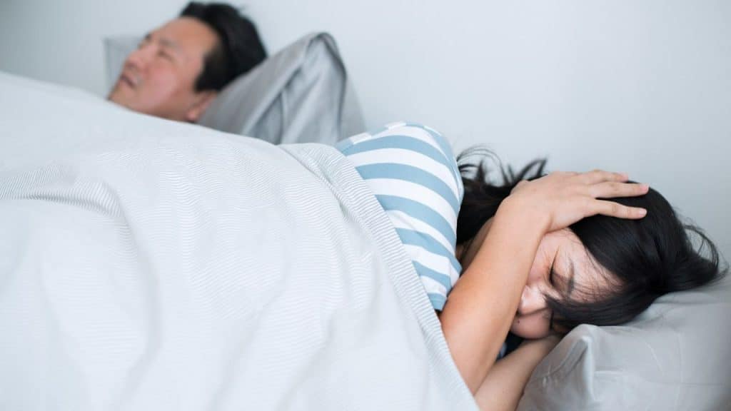 A woman lies in bed with her hand over her ear, looking frustrated, while a man sleeps soundly behind her.
