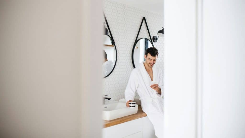 A man in a white robe is leaning against a bathroom counter, smiling at his phone.