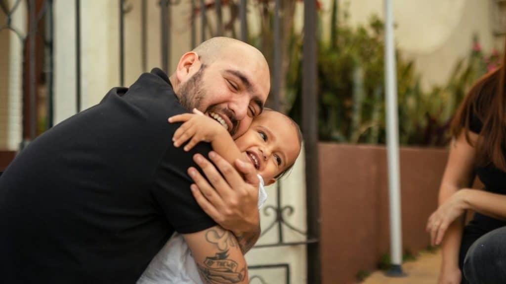 A man with tattoos on his arms is embracing a laughing toddler.