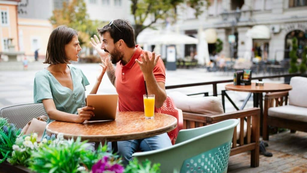 A man and a woman argue at an outdoor cafe table with a tablet.
