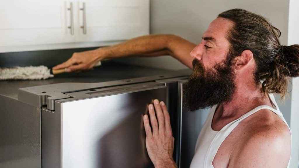 A man with a beard dusts the top of a refrigerator with a brush.