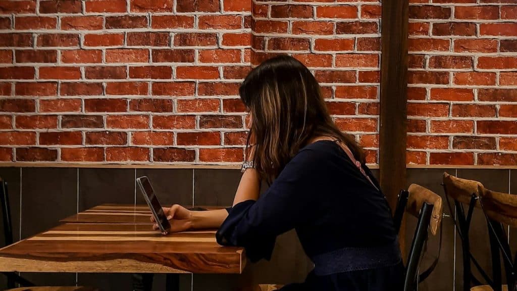 A woman with her head in her hand sits alone at a table, looking at her phone.