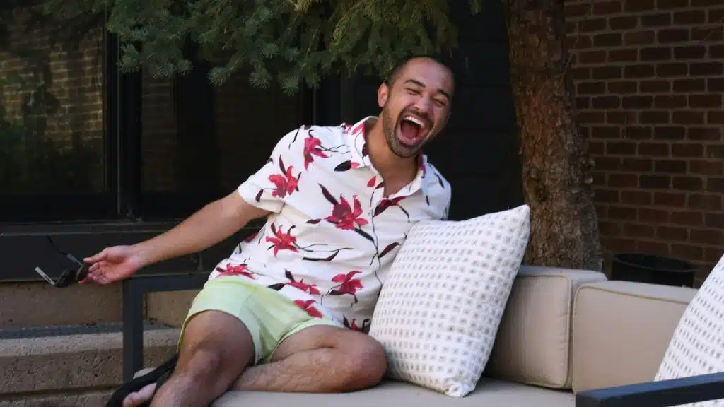 A man in a floral shirt and shorts laughs on an outdoor couch.