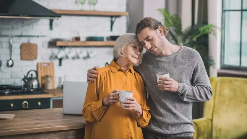 An older woman and younger man embrace in a kitchen, holding mugs.