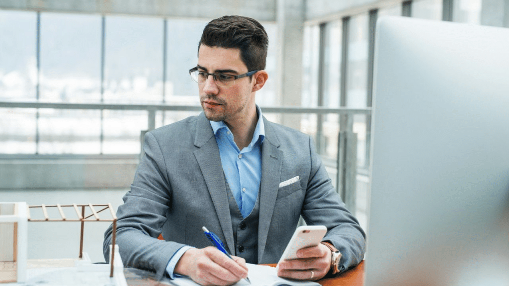 A man in a gray suit sits at a desk, looking at his phone and taking notes in a notebook.