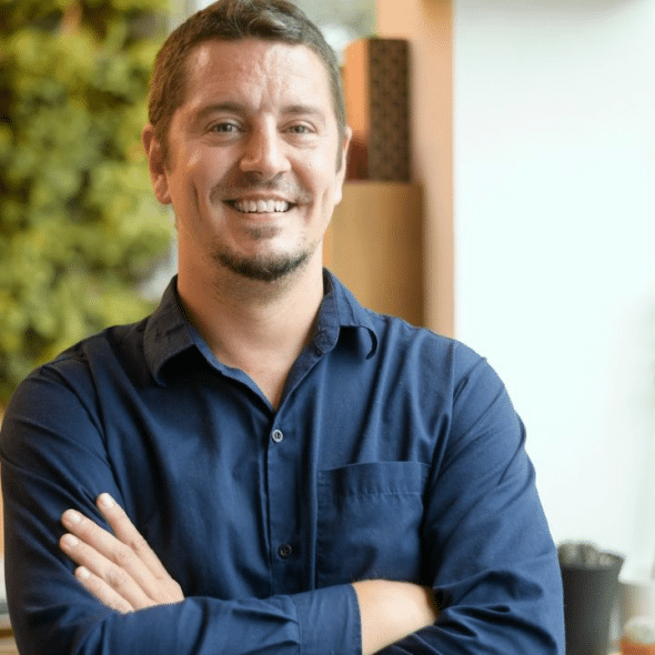 A man in a blue button-up shirt smiles at the camera with his arms crossed.