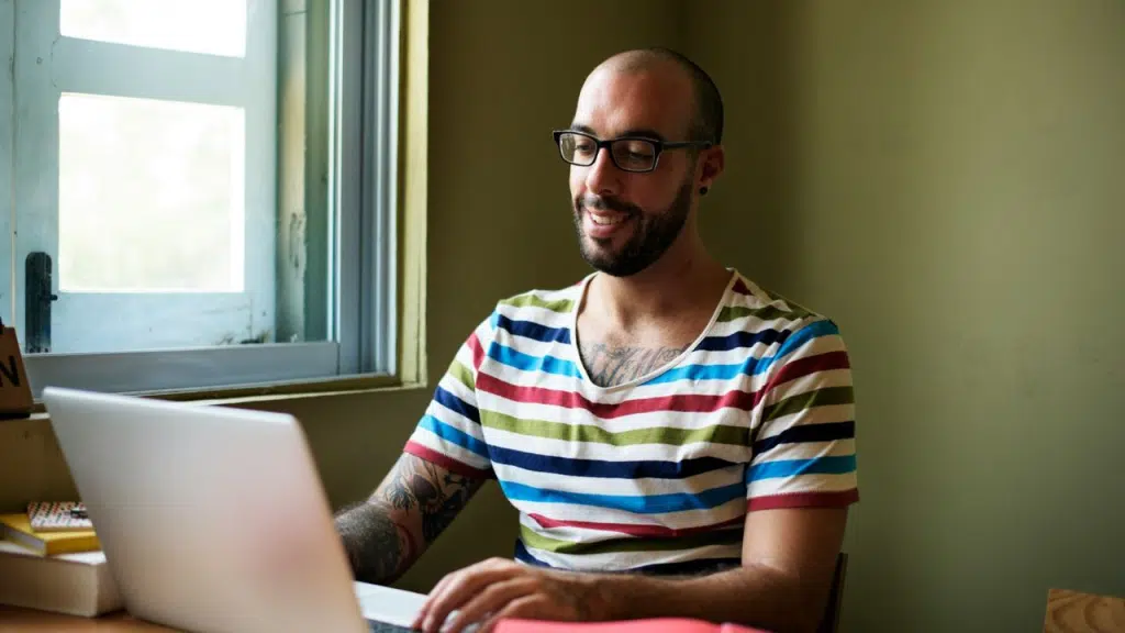 A man with glasses, a beard, and a striped shirt is smiling while looking at a laptop computer.