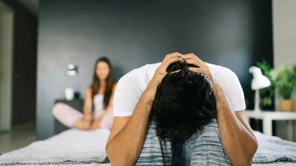 A man sitting on a bed with his head in his hands while a woman sits in the background.