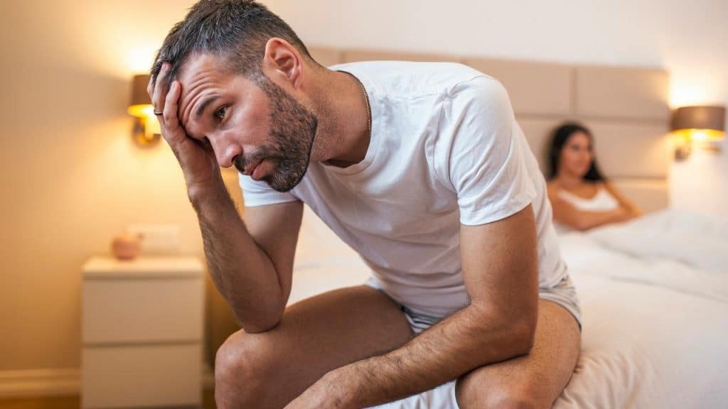 A man sitting on the edge of a bed holding his head while a woman sits in the background.