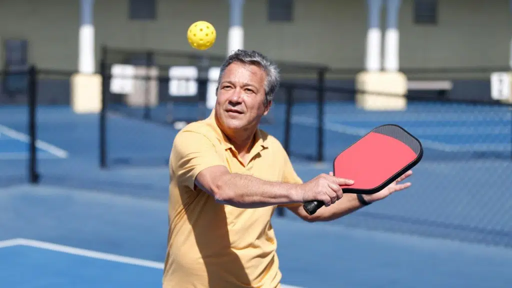 A man with gray hair in a yellow collared shirt plays pickleball on a blue court.