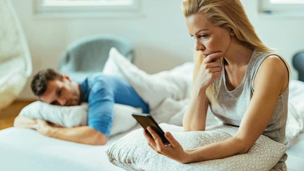 A woman sitting on a bed looking at her phone while a man lies in the background.