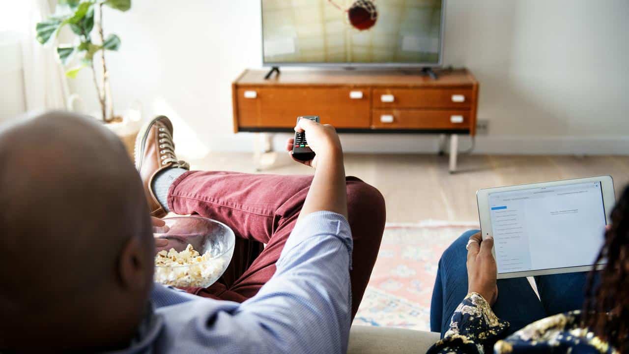 A couple watching TV with popcorn and a tablet.