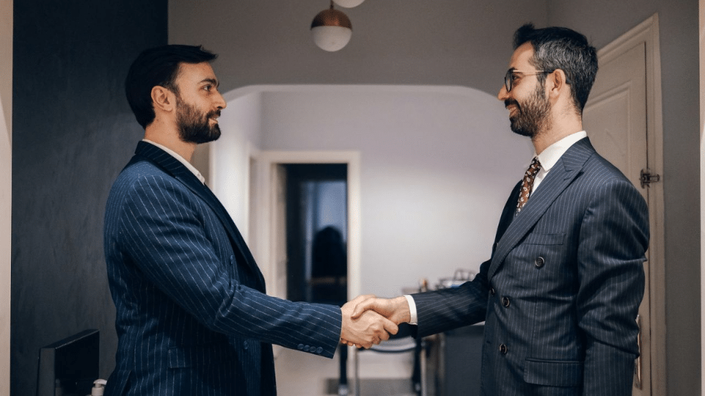Two men in pinstripe suits shake hands indoors while facing each other and smiling.