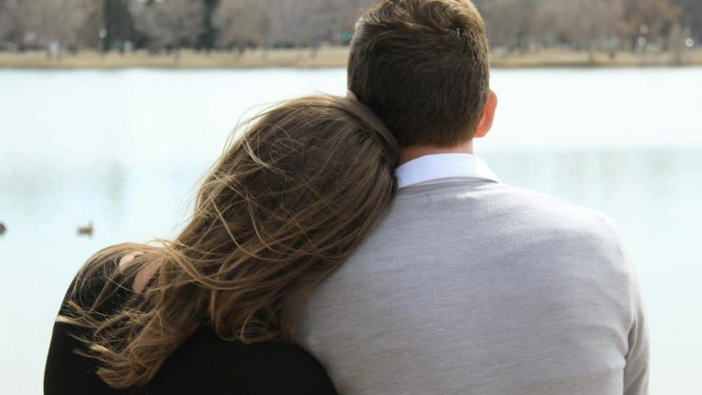 A couple sitting by the water with the woman resting her head on the man's shoulder.