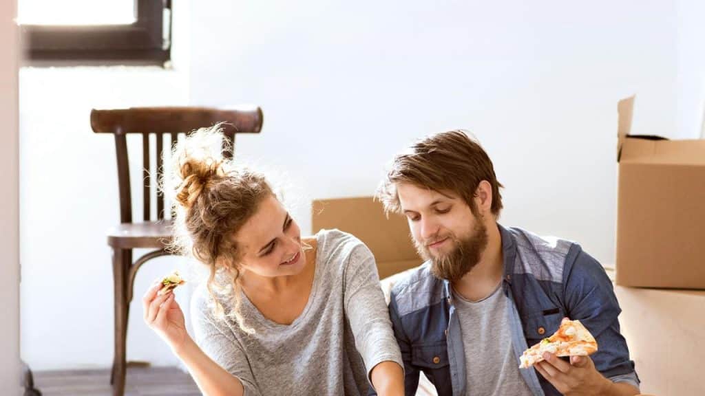 A couple sitting on the floor eating pizza with moving boxes around them.