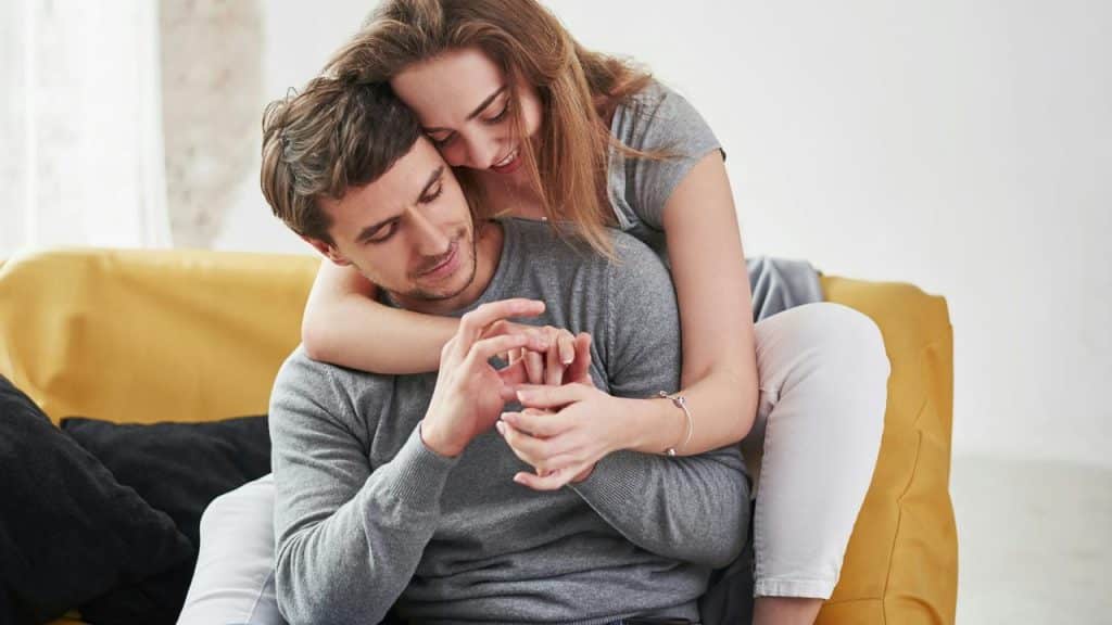 A woman hugging a man from behind while they smile and hold hands.