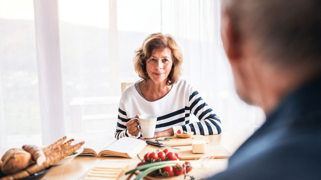 A mature woman holding a mug sits at a table and looks at a man.