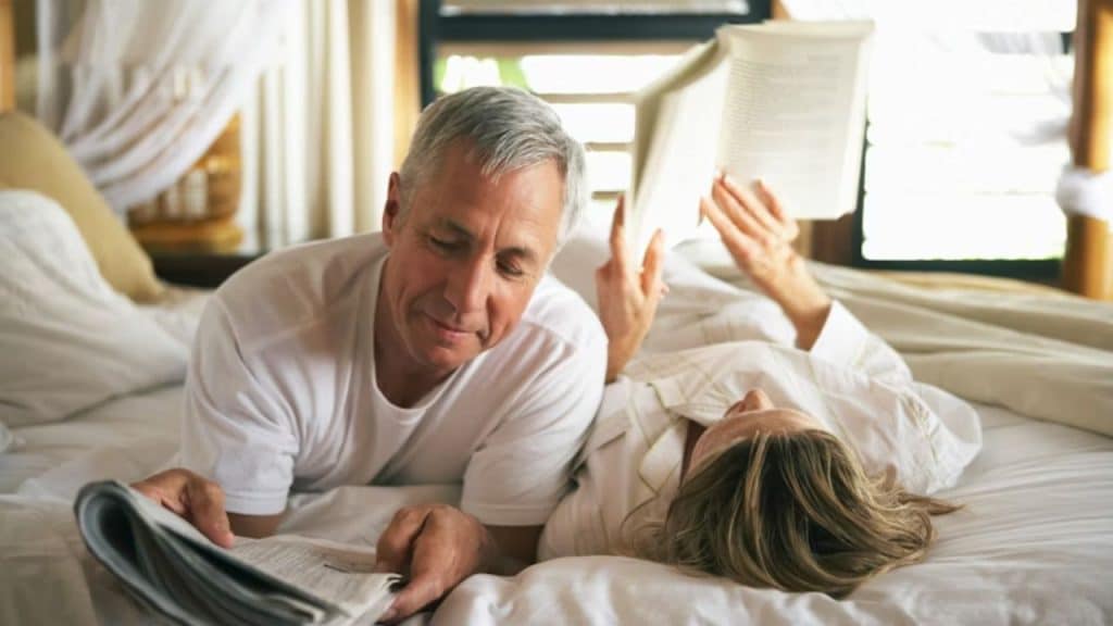 A man reading a newspaper while his wife reads a book on the bed